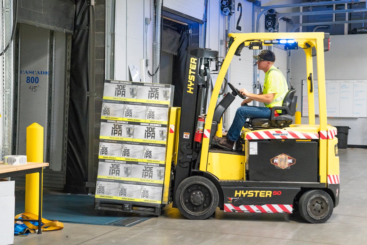 about-img-01 A worker drives a Hyster forklift moving Joe IPA boxes in a warehouse.